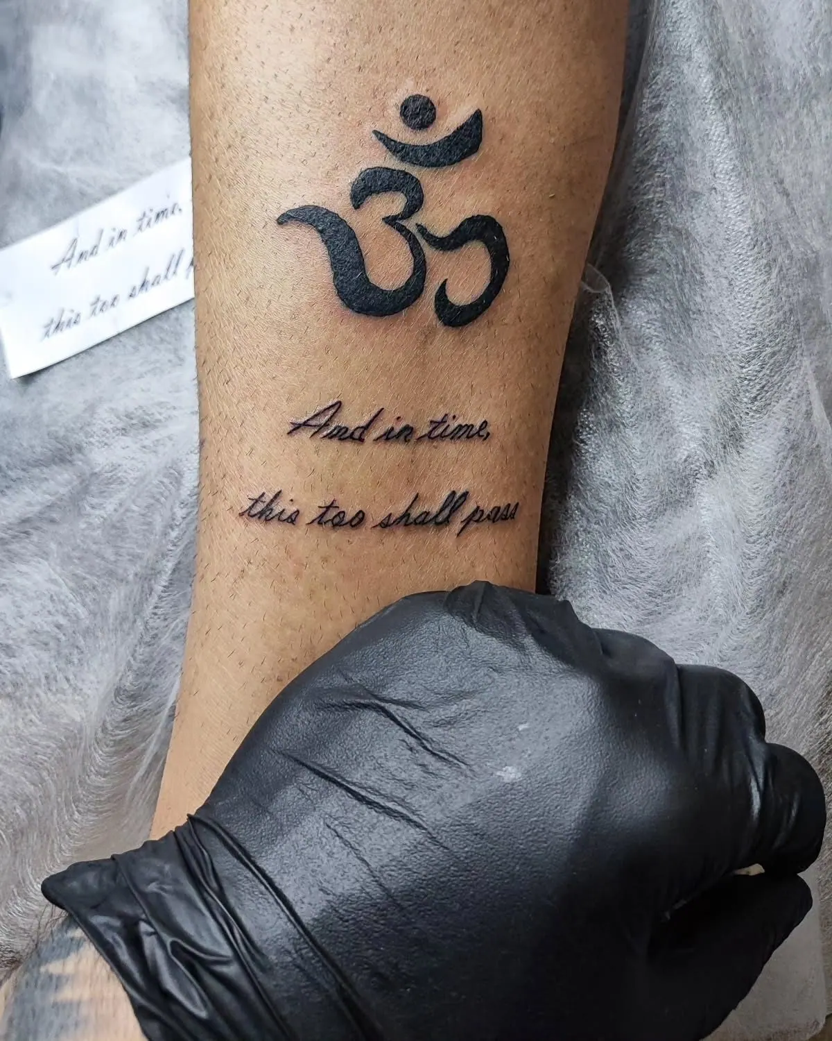 Om-symbool met daaronder de tekst 'And in time this too shall pass' op de onderarm