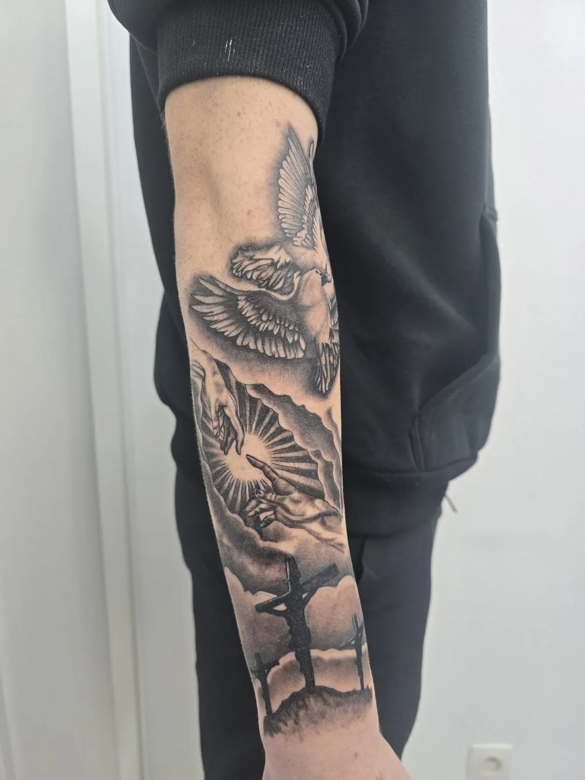 Zwart-grijze religieuze tattoo op de onderarm met een duif, twee reikende handen en de kruisiging