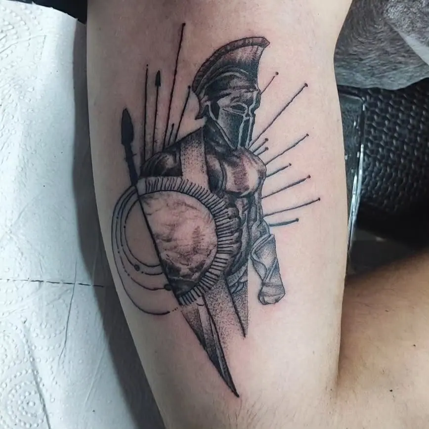 Zwart-grijze tattoo van een Spartaanse krijger met helm, schild en speer op de arm