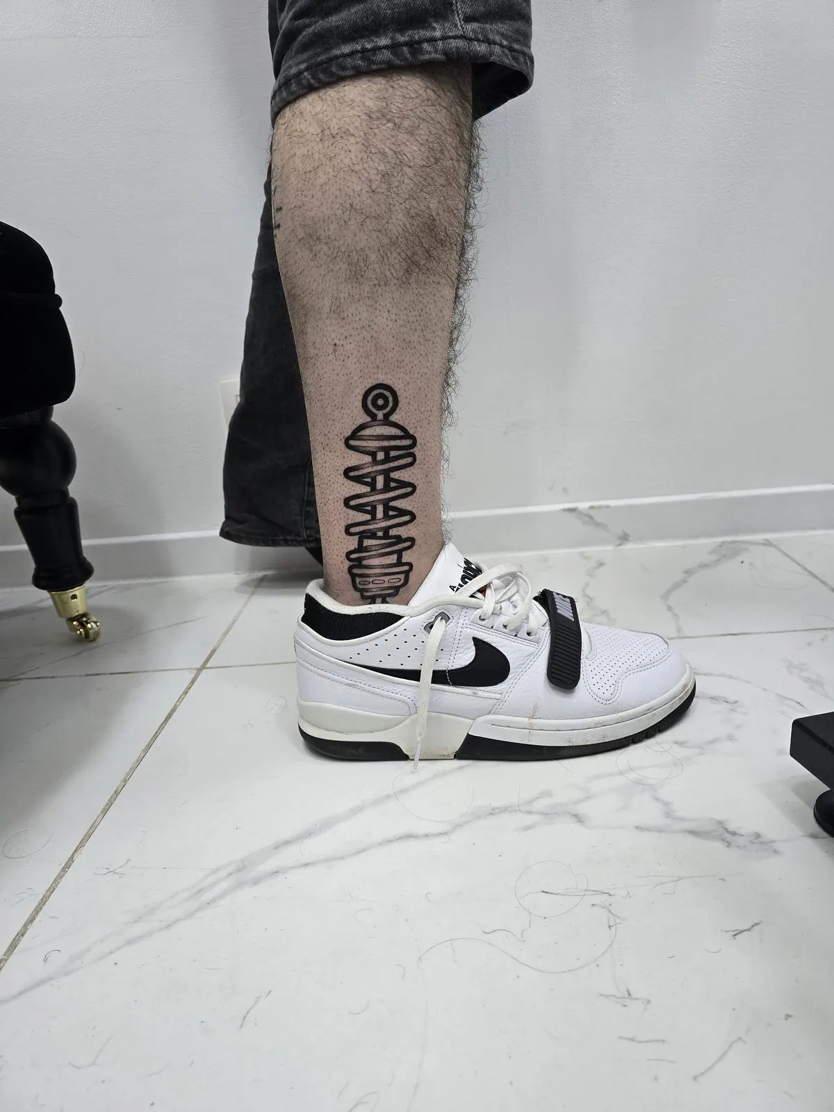 Minimalistische zwarte lijntattoo van een Tesla-spoel op het onderbeen